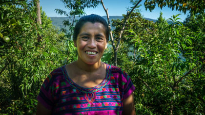 A woman coffee farmer in bright clothes stands in a forest and smiles at the camera.